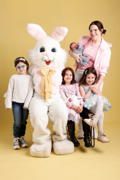 Mom with newborn and three young girls sitting on a bench with the Easter Bunny.  All are in front of a yellow backdrop.