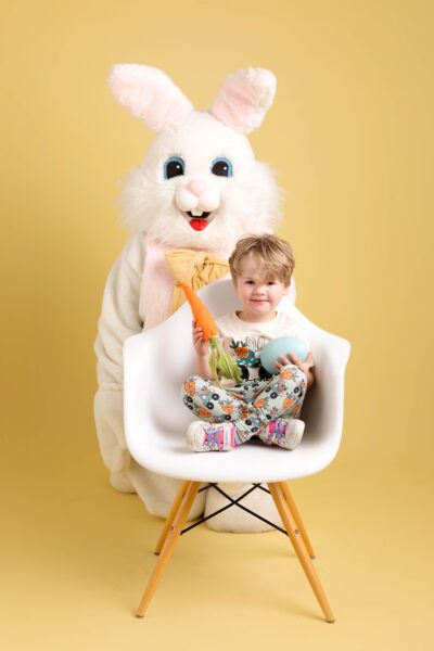 A toddler boy sits on a white chair in front of the Easter Bunny. He is holding a carrot and a large easter egg. They are on a yellow backdrop.