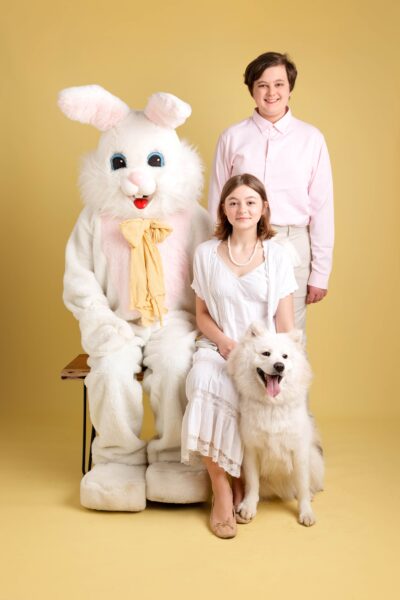 Teen boy and girl siblings sitting on a bench with the Easter Bunny. Their white dog sits next to them. All are in front of a yellow backdrop.